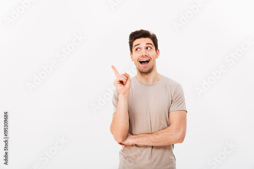Positive brunette man 30s wearing beige t-shirt gesturing finger aside on copyspace, isolated over white background