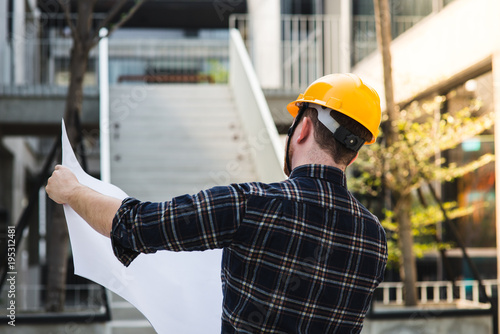 Back of engineer holding blueprint with building background.