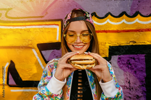 Attractive young woman, smiling cheerfully, holds tasty burger in two hands. Dressed in colorful jacket and cap, in sunglasses. Outdoors, near the wall with graffiti.