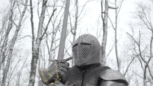 Round view of brave warrior wearing steel armor and helmet preparing for swordfight, on battlefield in winter snowy wood slow motion. Reconstruction of historical times