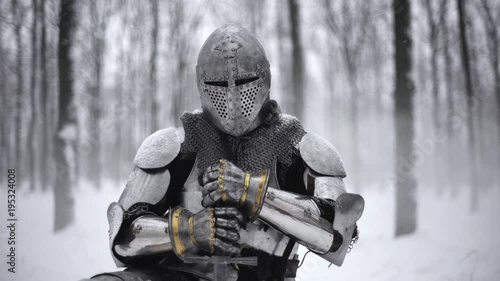 Portrait of medieval knight in helmet holding steel sword and kneeling on winter nature, in slow motion. Fantasy and reenactment