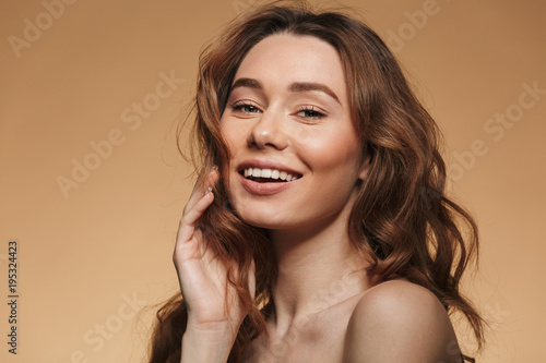 Feminine pleased girl 20s with long curly brown hair looking at camera with sincere smile, isolated over beige background
