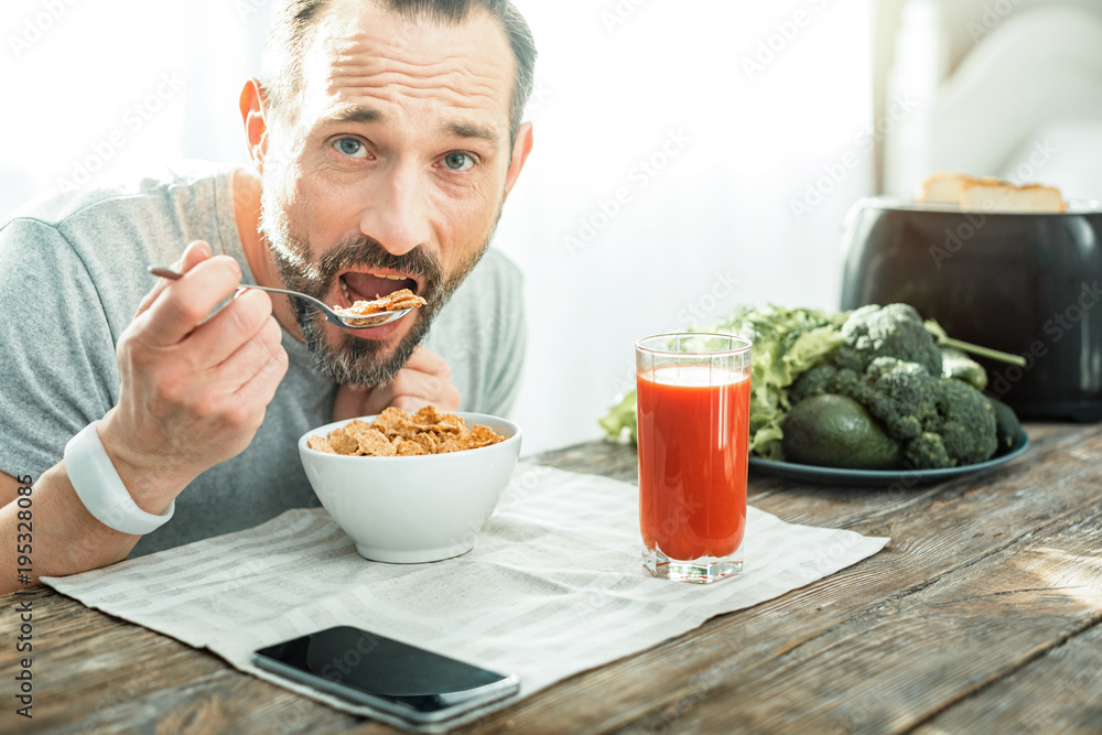 Healthy food. Handsome pleasant beardful man sitting in the bright kitchen holding a spoon and eating flakes. Healthy food. Handsome pleasant beardful man sitting in the bright kitchen holding a spoon and eating flakes.