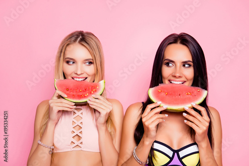 Portrait of hot, comic, childish, cute, seductive, blonde, brunette tourists, ladies in swim suits looking to each other with eye, having lobes of watermelon in hands, isolated on pink background