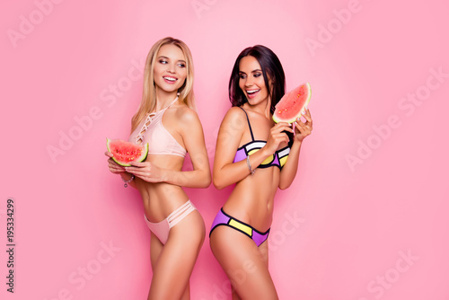 Charming, hot, pretty, seductive, positive, trendy, stylish ladies, tourists in swimsuits having pieces of watermelon in hands, standing back to back looking to each other over pink background