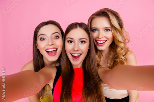 Self portrait of charming, funky, nice, attractive, pretty girls making selfie on smart phone over pink background, with open mouths, laughing, screaming, shouting, glad, happiness