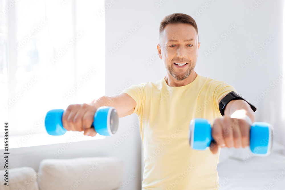 Key trend. Jovial jolly handsome man using dumbbells while exercising and looking at hand Key trend. Jovial jolly handsome man using dumbbells while exercising and looking at hand