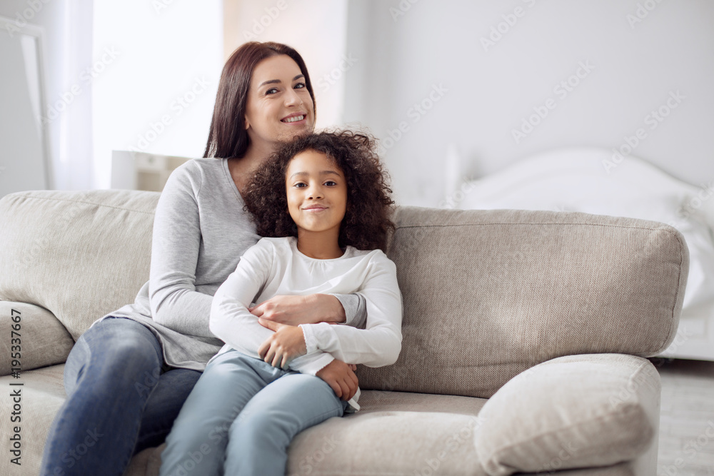 I feel happy. Beautiful cheerful young dark-haired woman smiling and hugging her daughter while sitting on the couch I feel happy. Beautiful cheerful young dark-haired woman smiling and hugging her daughter while sitting on the couch