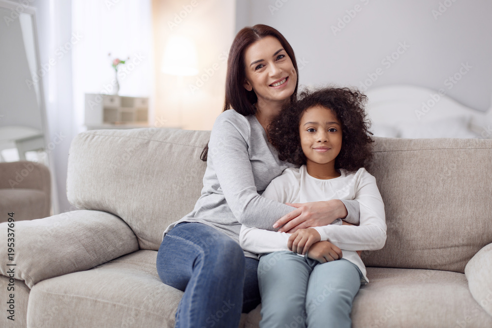 My dear daughter. Beautiful content young dark-haired woman smiling and hugging her daughter while sitting on the couch My dear daughter. Beautiful content young dark-haired woman smiling and hugging her daughter while sitting on the couch