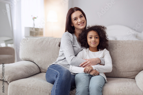 My dear daughter. Beautiful content young dark-haired woman smiling and hugging her daughter while sitting on the couch
