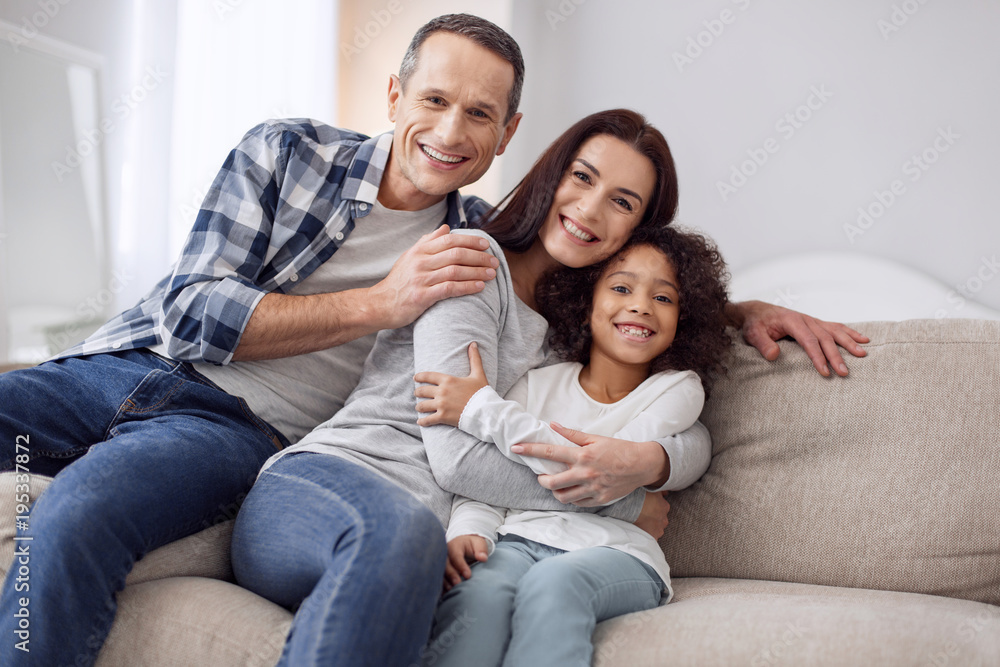 Happy together. Beautiful content curly-haired girl smiling and sitting on the couch with her parents and they hugging each other Happy together. Beautiful content curly-haired girl smiling and sitting on the couch with her parents and they hugging each other