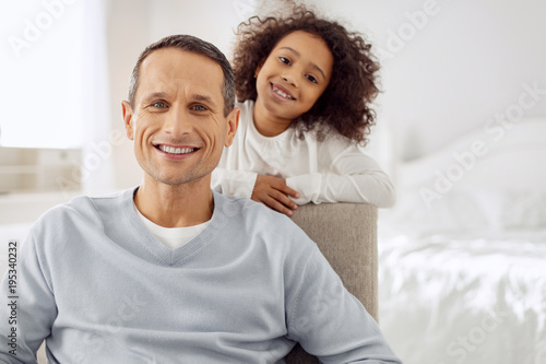Happy fatherhood. Attractive alert dark-haired father sitting in the arm-chair and smiling and his daughter standing behind him