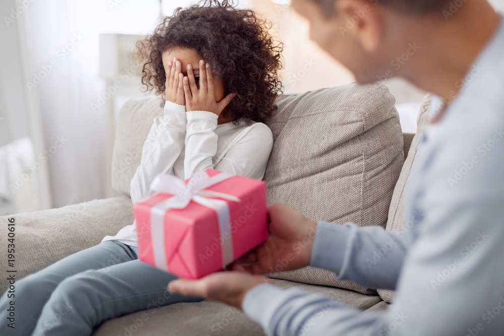 My gift. Inspired lovely curly-haired girl sitting on the sofa and closing her and her daddy holding a gift My gift. Inspired lovely curly-haired girl sitting on the sofa and closing her and her daddy holding a gift