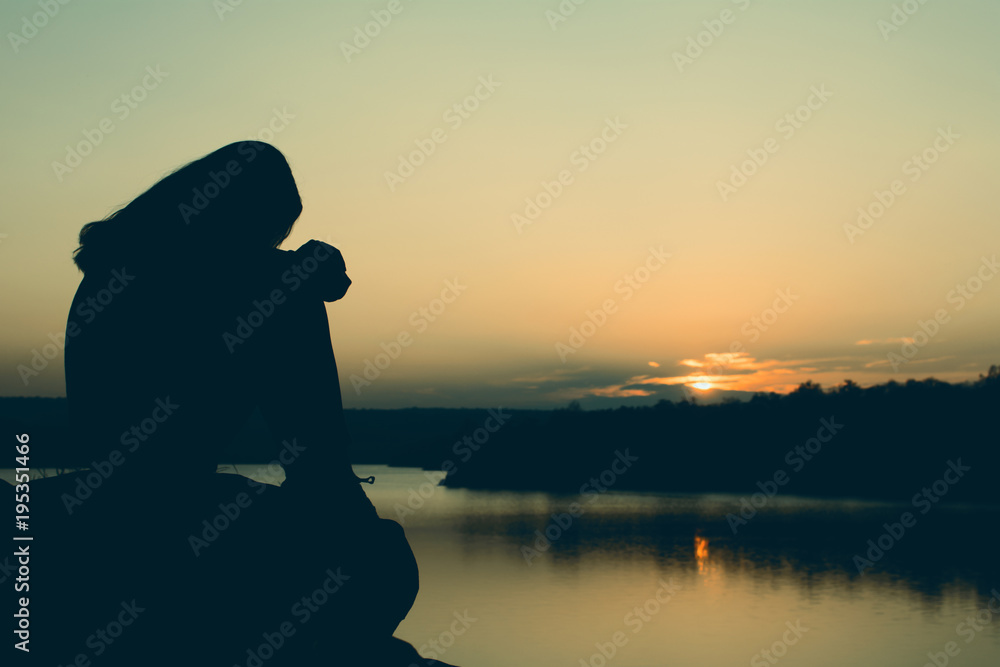 Silhouette of anxious woman sitting on the rock in nature during sunset