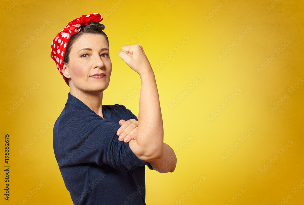 Fototapeta premium Self-confident middle aged woman with a clenched fist rolling up her sleeve, text space, tribute to american icon Rosie Riveter