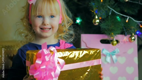 Close-up of a petite girl of three years standing near a Christmas tree and holding a gift box in her hand.