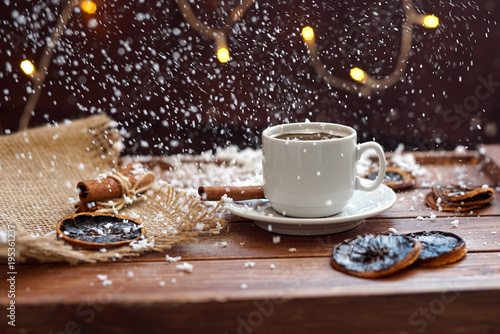 Чашка кофе с салфеткой из мешковины, сушеными дольками лимона и палочками корицы на деревянном коричневом фоне с огнями гирлянды под падающим снегом