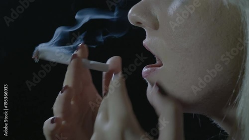 Close up of a depressed blonde teenager girl sitting in the dark lightning a weed cigarette inhaling and exhaling clouds of smoke