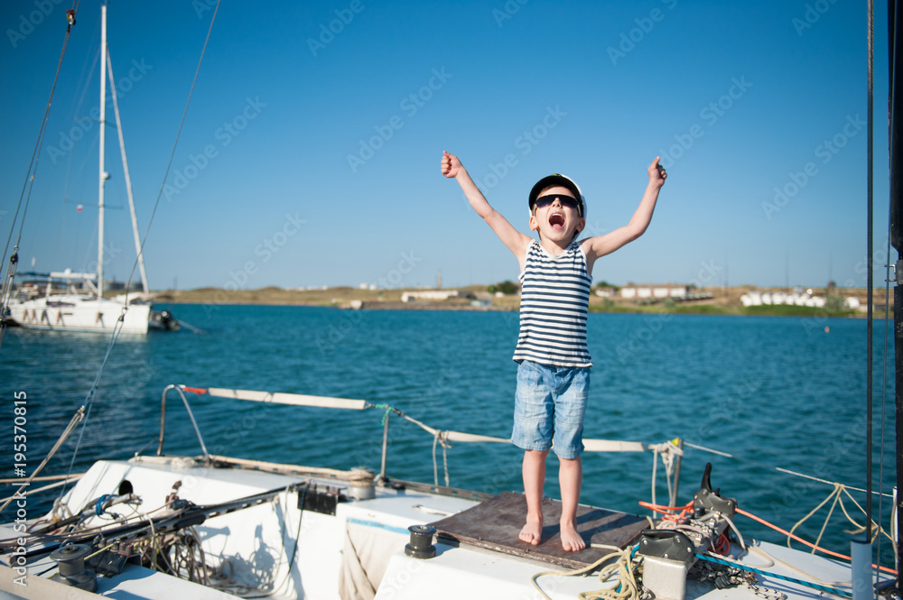 cute little boy in captain hat and sunglasses on yacht screams joyfully waving his hands