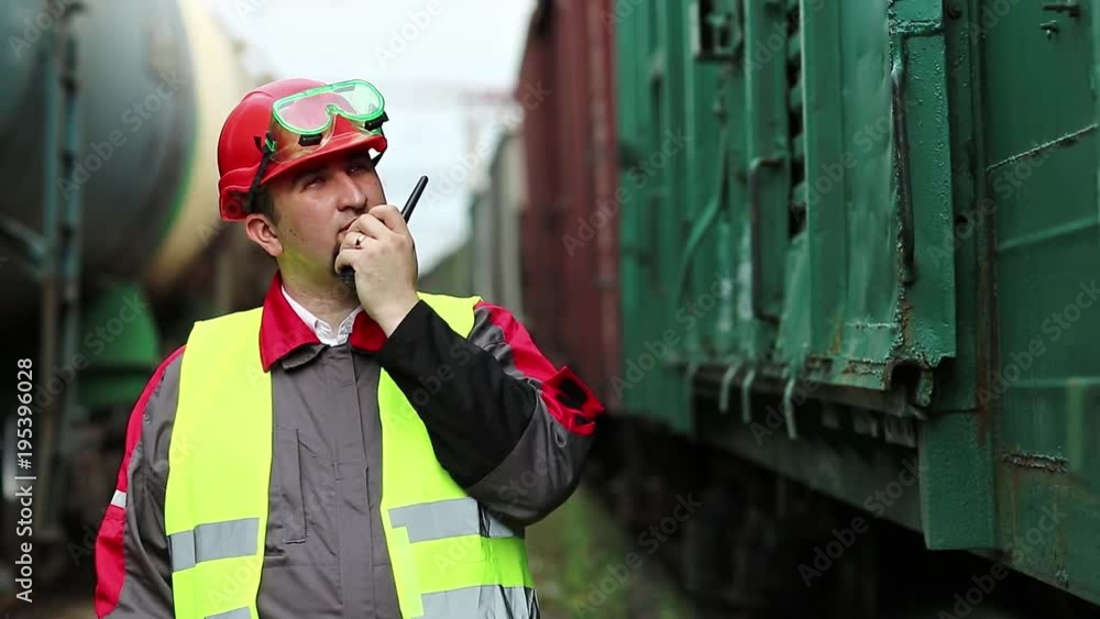 Railway employee on railway line near freight station speaks via portable radio transmitter, worker communicates via radio station. Railroader with portable radio transmitter near goods trains Railway employee on railway line near freight station speaks via portable radio transmitter, worker communicates via radio station. Railroader with portable radio transmitter near goods trains