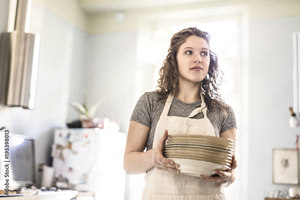 young woman prepares the table by carrying a stack of dishes in a bright kitchen young woman prepares the table by carrying a stack of dishes in a bright kitchen