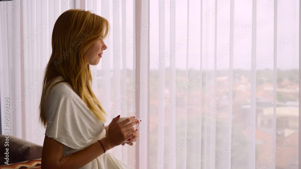 Young caucasian woman in dress with white cup standing near big panoramic window, looking out and at the camera, then drinking beverage Young caucasian woman in dress with white cup standing near big panoramic window, looking out and at the camera, then drinking beverage