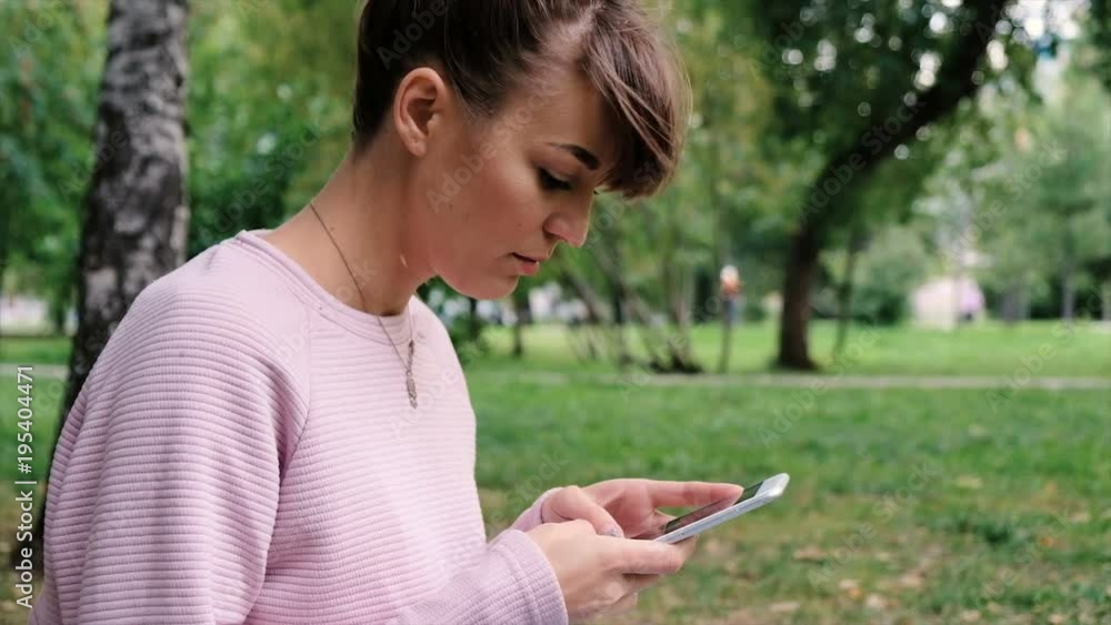Portrait of relaxed young lady in a summer park reading a text message on her mobile phone. Beautiful young girl in pink sweatshirt using her cell phone and looking at the camera, outdoor. Portrait of relaxed young lady in a summer park reading a text message on her mobile phone. Beautiful young girl in pink sweatshirt using her cell phone and looking at the camera, outdoor.