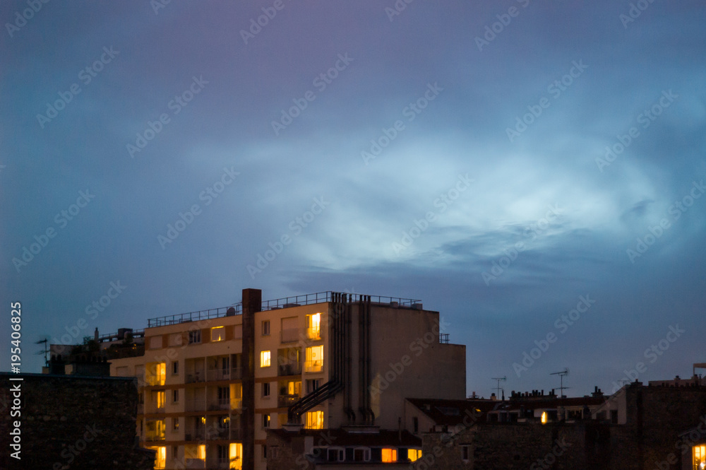 Night is falling on a residential building with illuminated rooms in a popular district under a dark blue sky.