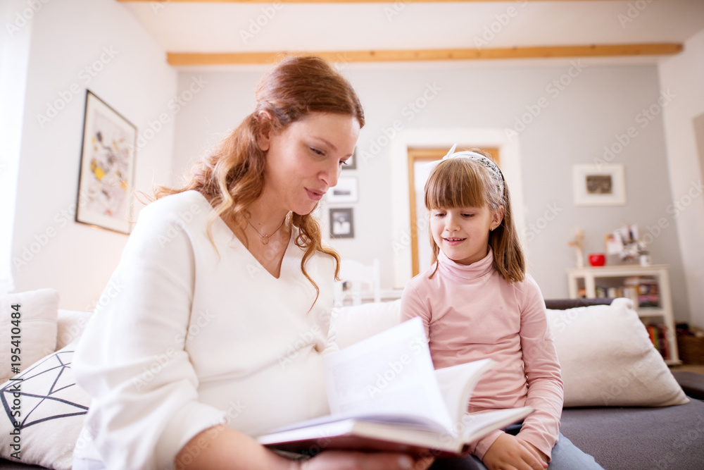 Sweet toddler girl sitting next to her careful pregnant mum and reading the book together. Sweet toddler girl sitting next to her careful pregnant mum and reading the book together.