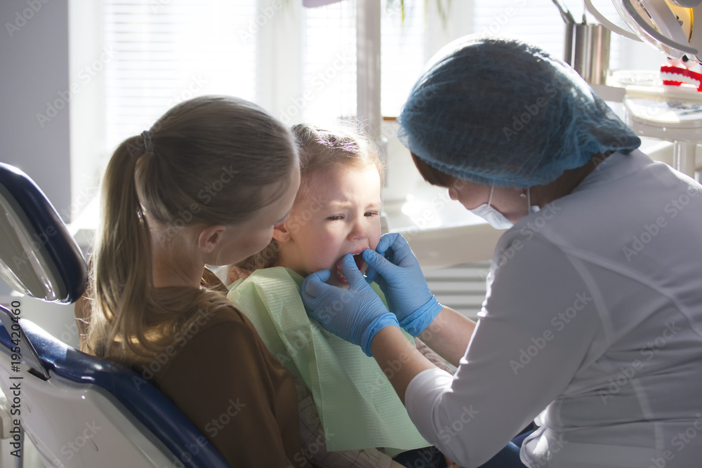 Girl, her mom and the dentist in the dental office, the stomatologist examining and consulting, daughter playing and complaining Girl, her mom and the dentist in the dental office, the stomatologist examining and consulting, daughter playing and complaining