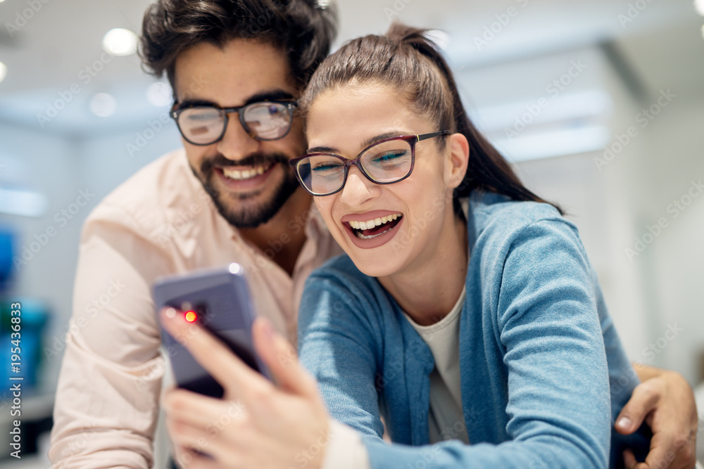 Young satisfied stylish charming couple testing the new model of a mobile from the desk in the tech store. Young satisfied stylish charming couple testing the new model of a mobile from the desk in the tech store.
