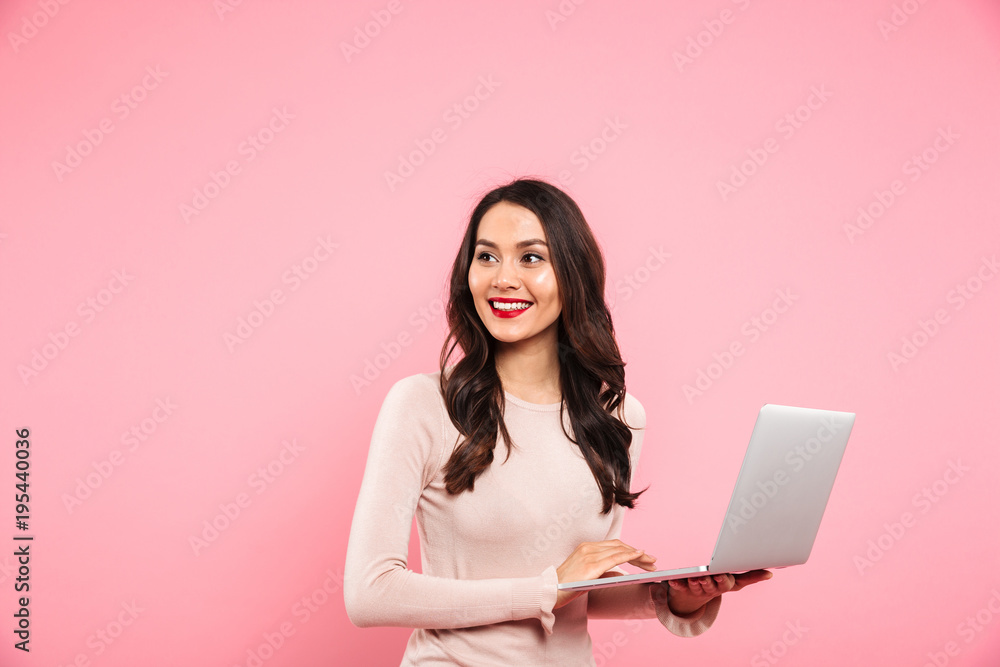 Photo of brunette cute woman with long hair holding silver laptop and looking aside on copyspace with candid smile, isolated over pink background Photo of brunette cute woman with long hair holding silver laptop and looking aside on copyspace with candid smile, isolated over pink background