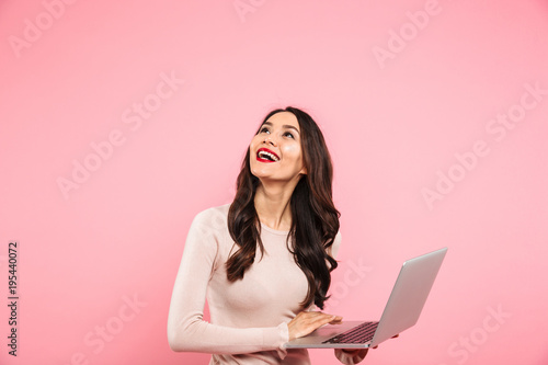 Photo of brunette cute woman in casual sweater holding silver notebook and looking upward on copyspace with candid smile, isolated over pink background