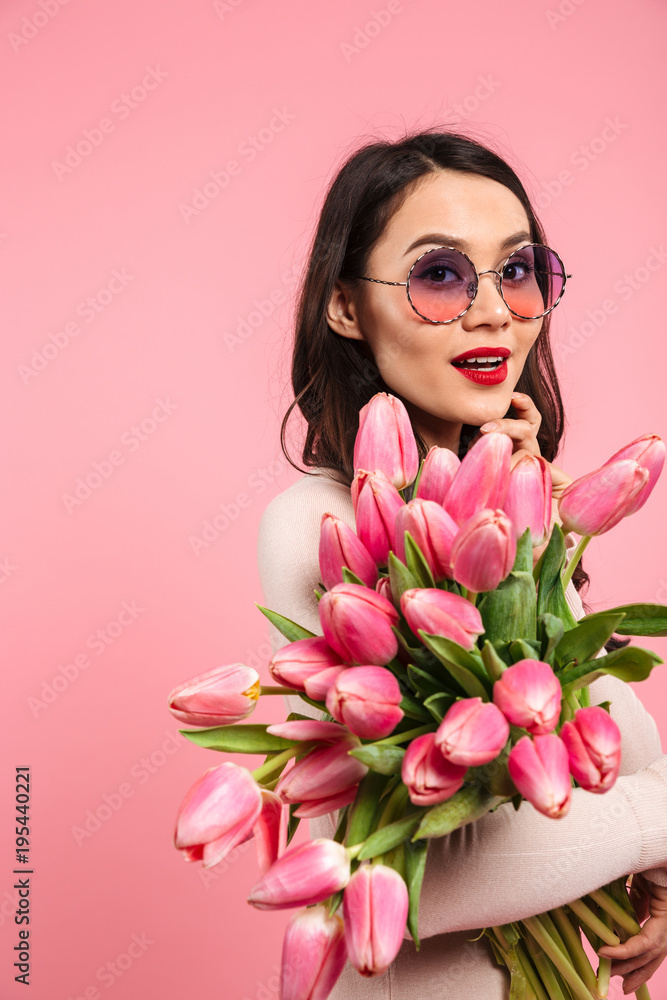 Photo of attractive woman with long dark hair in fashionable sunglasses posing on camera with lots of spring flowers, isolated over pink background Photo of attractive woman with long dark hair in fashionable sunglasses posing on camera with lots of spring flowers, isolated over pink background