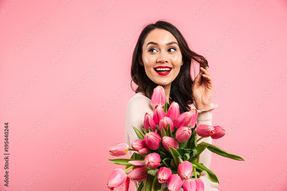 Attractive woman with long dark hair posing on camera with lots of spring flowers and looking aside, isolated over pink background