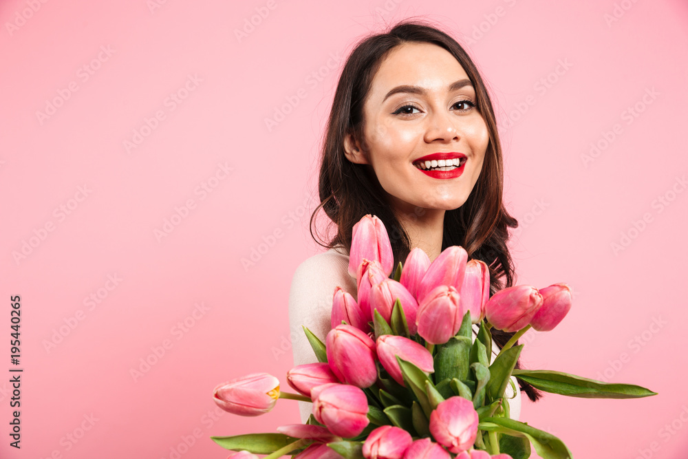 Wonderful woman 20s with beautiful lovely look posing on camera holding bunch of spring tulips, isolated over pink background Wonderful woman 20s with beautiful lovely look posing on camera holding bunch of spring tulips, isolated over pink background