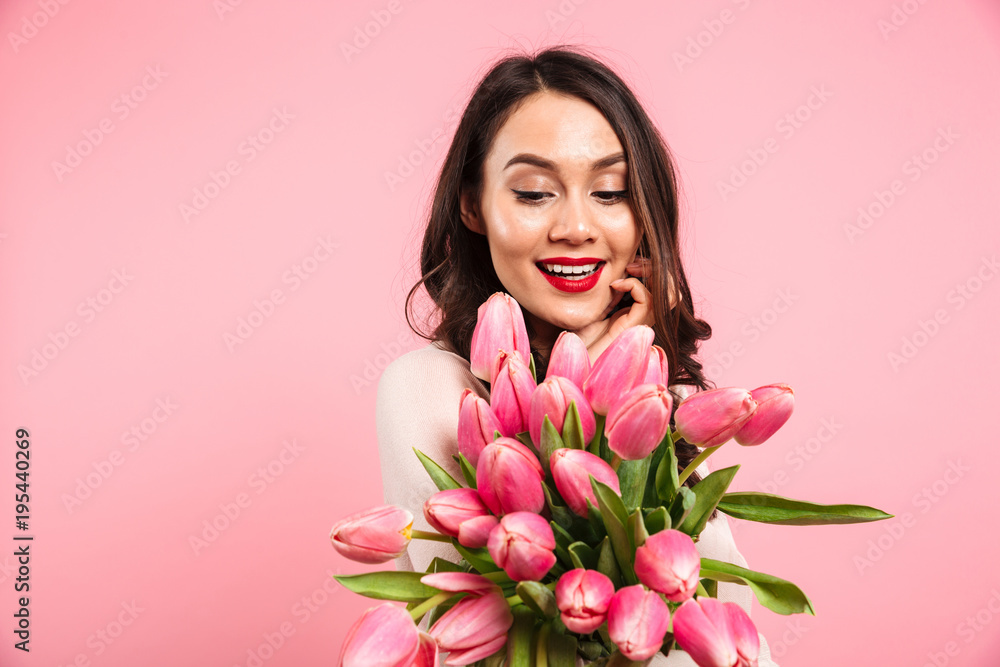 Magnificent picture of woman with beautiful smile posing on camera with lots of spring flowers, isolated over pink background