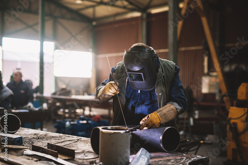 Welder in protective uniform and mask welding metal pipe on the industrial table while sparks flying.