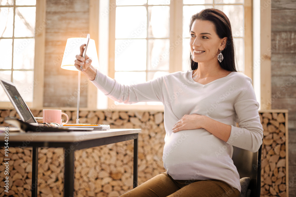 Smile on photo. Jovial attractive pregnant woman taking photo while grinning and putting hand on belly Smile on photo. Jovial attractive pregnant woman taking photo while grinning and putting hand on belly
