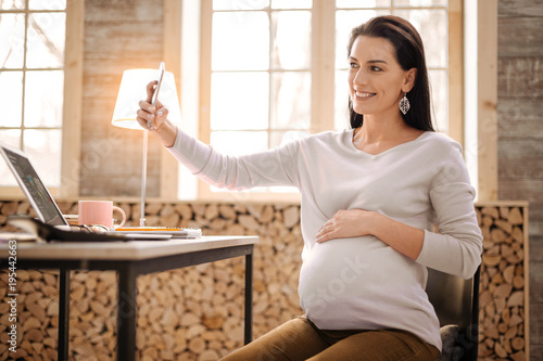 Smile on photo. Jovial attractive pregnant woman taking photo while grinning and putting hand on belly