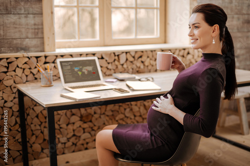 Herbal tea. Hopeful optimistic pregnant businesswoman caressing tummy while grinning and enjoying tea