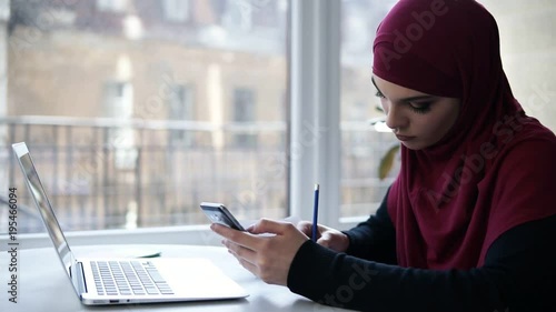 Young suposedly muslim girl with purple hijab on her head writes something down from her smartphone, sitting in a light indoor space with glass windows on the background photo