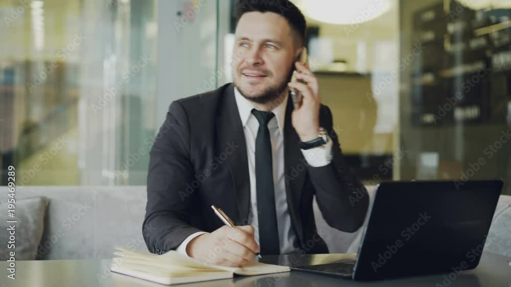 Happy Caucasian businessman sitting, smiling, talking on smartphone with partner and writing down information in notepad while looking at laptop in cafe Happy Caucasian businessman sitting, smiling, talking on smartphone with partner and writing down information in notepad while looking at laptop in cafe