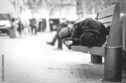 Poor homeless man or refugee sleeping on the wooden bench on the urban street in the city, social documentary concept, selective focus, black and white photo