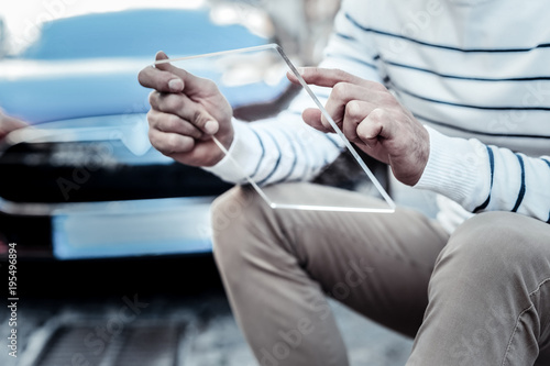 Technological progress. Close up of an innovative tablet being in hands of a cheerful positive man while sitting near his car