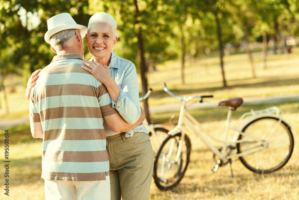Wonderful mood. Delighted nice positive woman looking at you and smiling while hugging her husband Wonderful mood. Delighted nice positive woman looking at you and smiling while hugging her husband