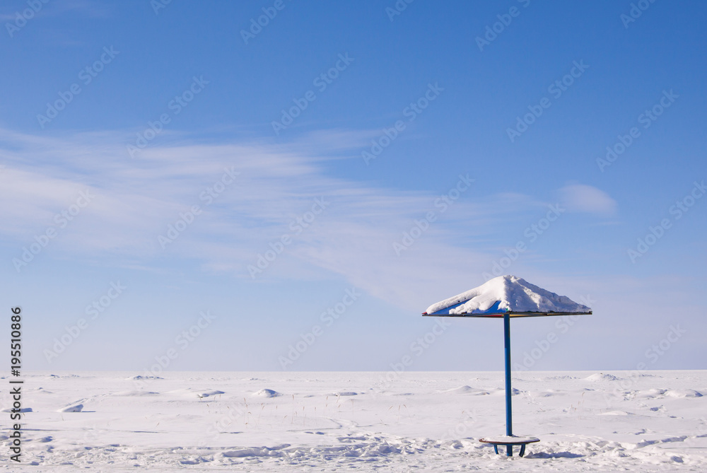 Scenic winter view of lonely sun umbrella on deserted beach covered by snow. Blue sky and frozen sea background.