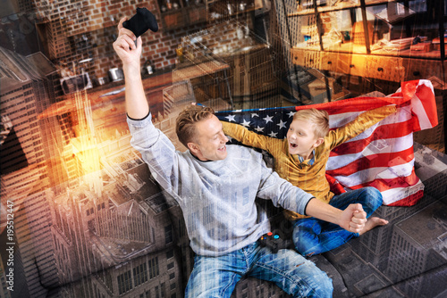 Winners. Cheerful emotional young man putting a hand with a game console up and celebrating the victory while his son sitting with the American flag and looking happy