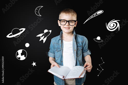 Curious boy. Clever cheerful curious boy wearing big glasses and feeling glad while holding a big notebook and reading the information about the space