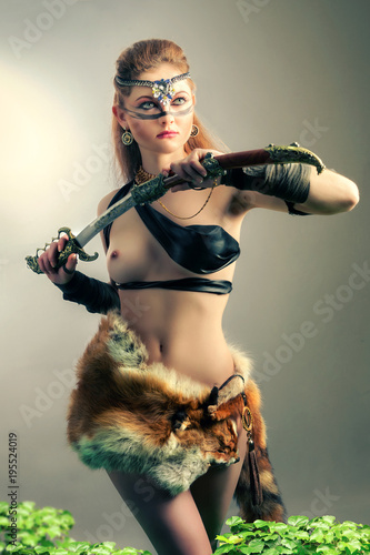 a pretty young woman with long red hair is holding a sword in her hands. She is only  wearing a mask and skins of wild animals . Her breast is perfect. She is standing in the grey background photo